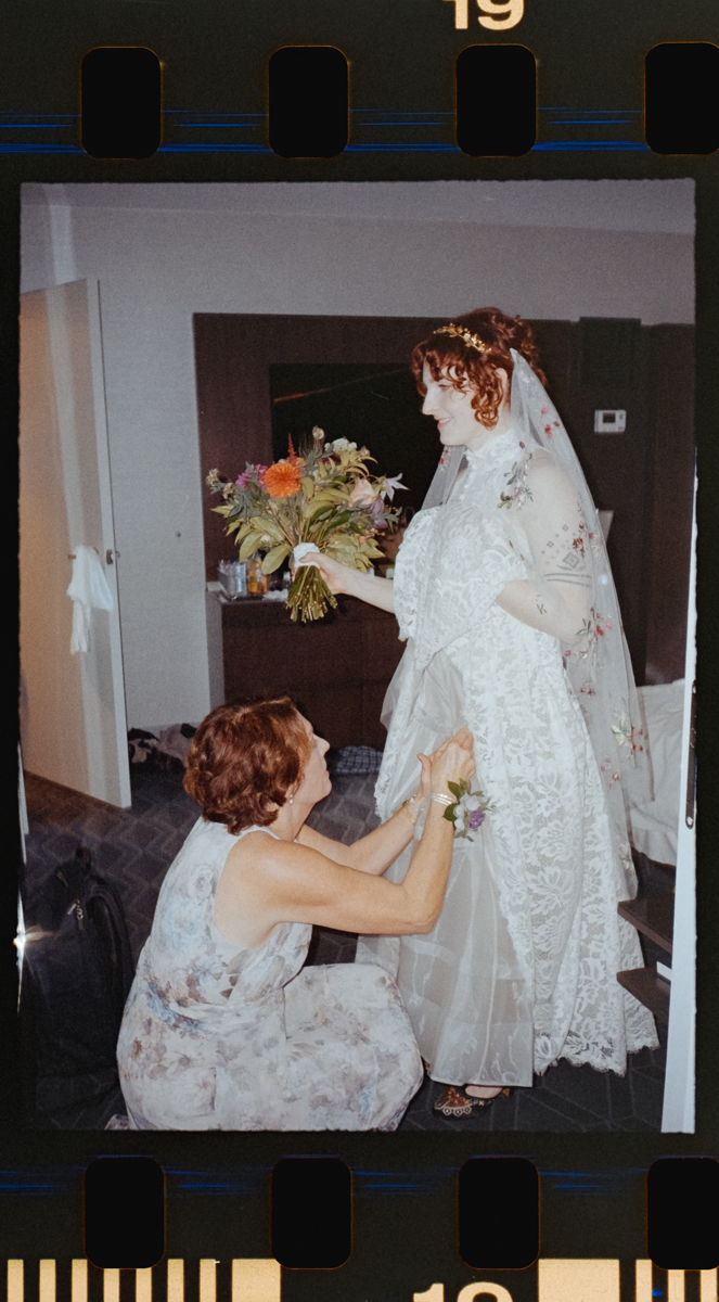 A woman in a floral dress kneels to adjust the lace dress of a bride holding a colorful bouquet. The bride, beneath a long veil, stands in a modern room—captured with vintage film style by Glass House Cambridge Wedding Photography.