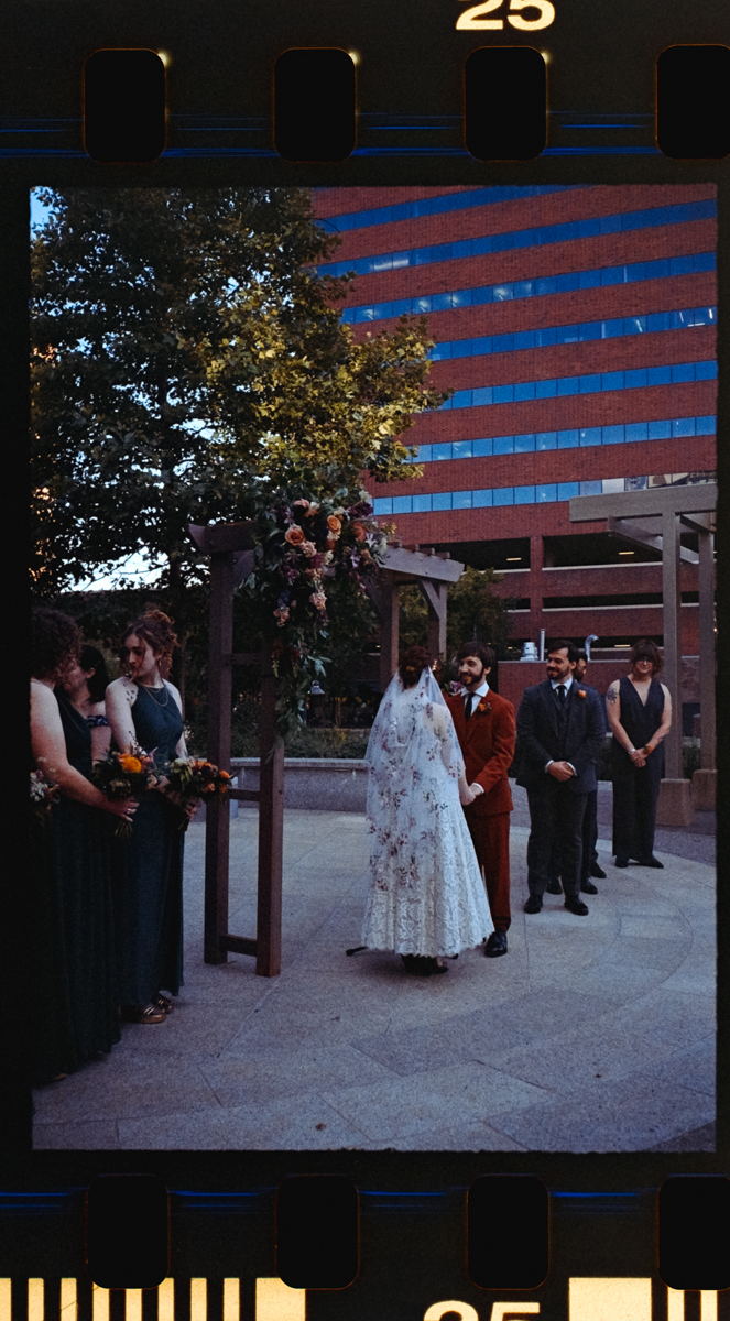 A couple exchanges vows during their wedding ceremony, surrounded by friends and family in an urban setting, with floral decorations and modern buildings in the background.