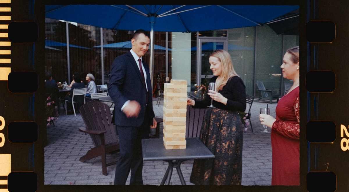 Three individuals engaged in a game of Jenga outdoors, with a casual atmosphere and drinks in hand.