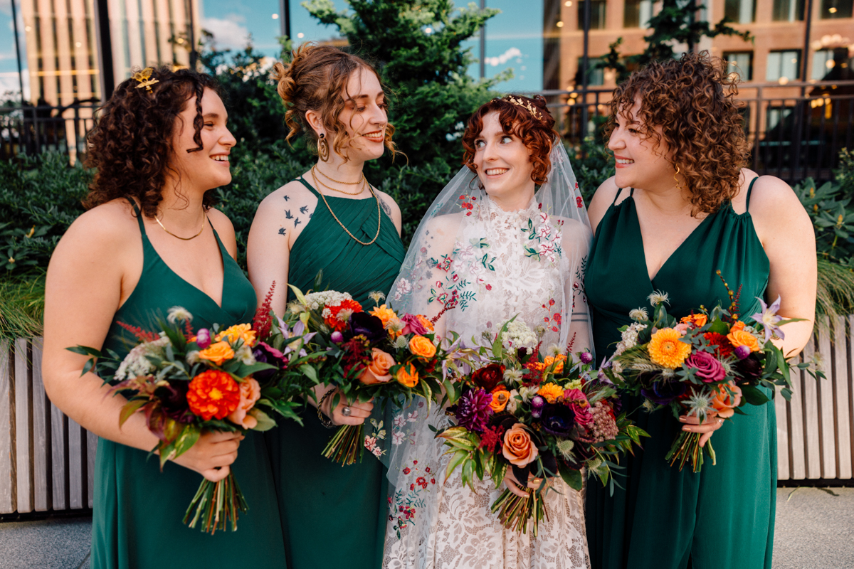 A joyful bridal party featuring four women in elegant green dresses, holding vibrant floral bouquets and smiling together outdoors.