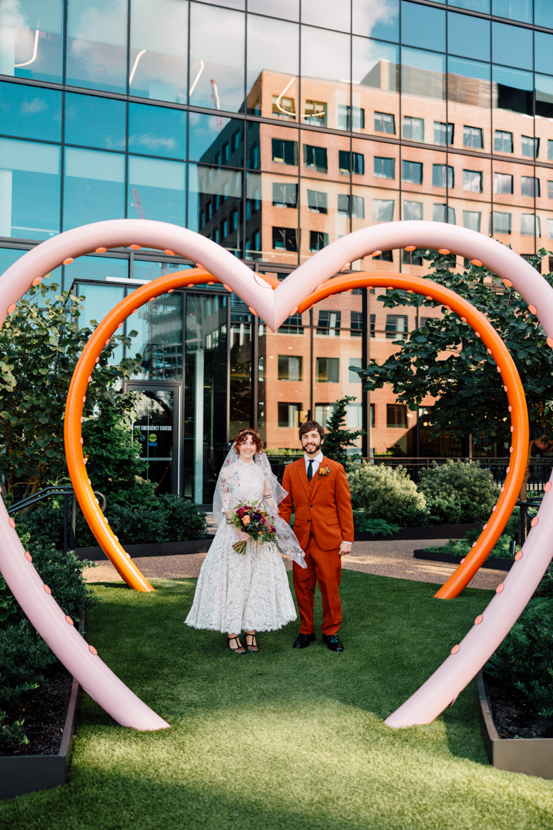 A happy couple stands beneath a large heart-shaped arch, surrounded by greenery, showcasing a vibrant wedding atmosphere.