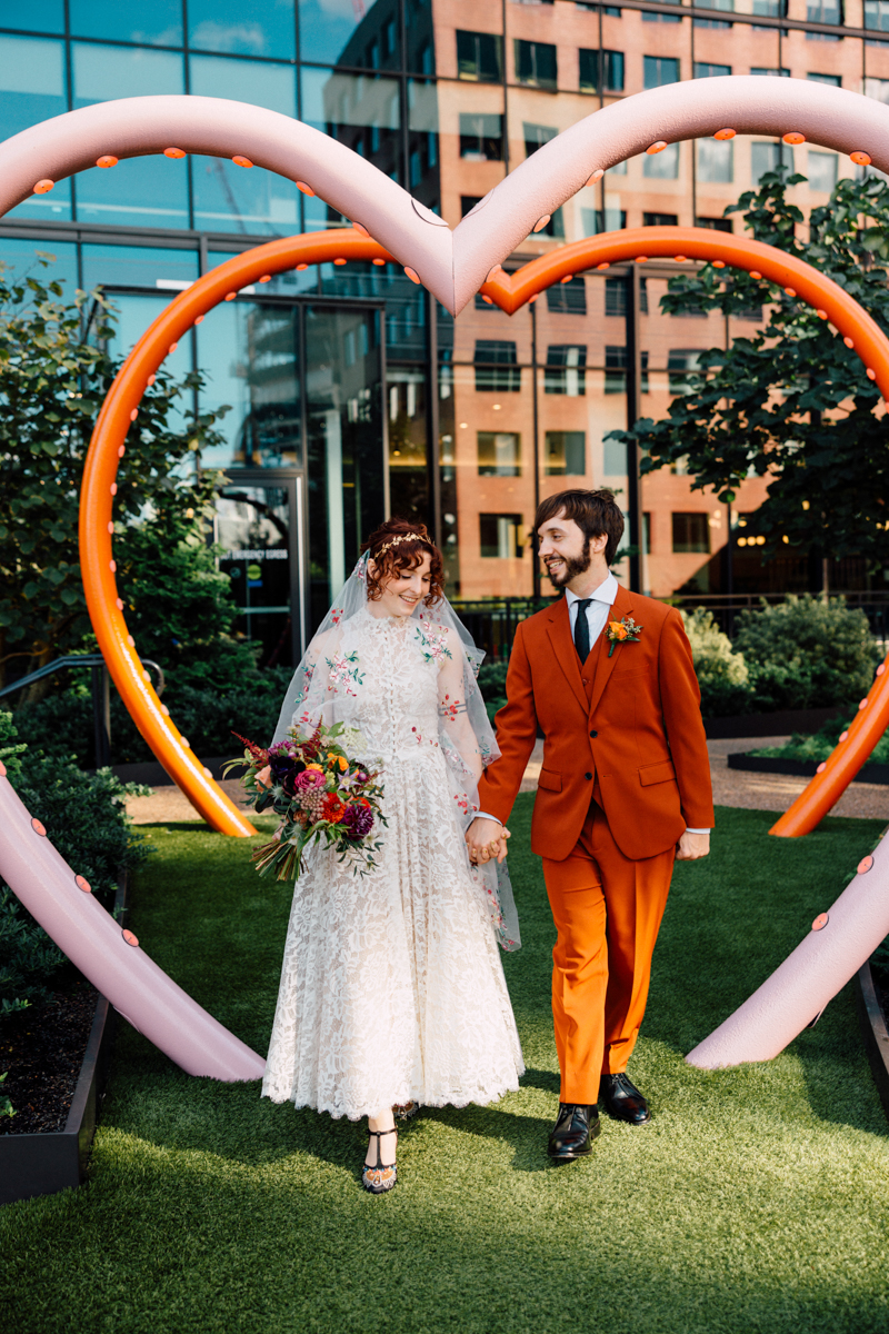A happy couple walks hand in hand under a colorful heart-shaped arch, surrounded by greenery and modern buildings, with the bride holding a vibrant bouquet and wearing a lace dress, while the groom sports an orange suit.