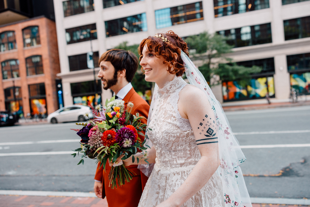 A joyful couple walks hand-in-hand in an urban setting, with the bride wearing a lace gown and carrying a vibrant bouquet, while the groom dons a stylish suit.