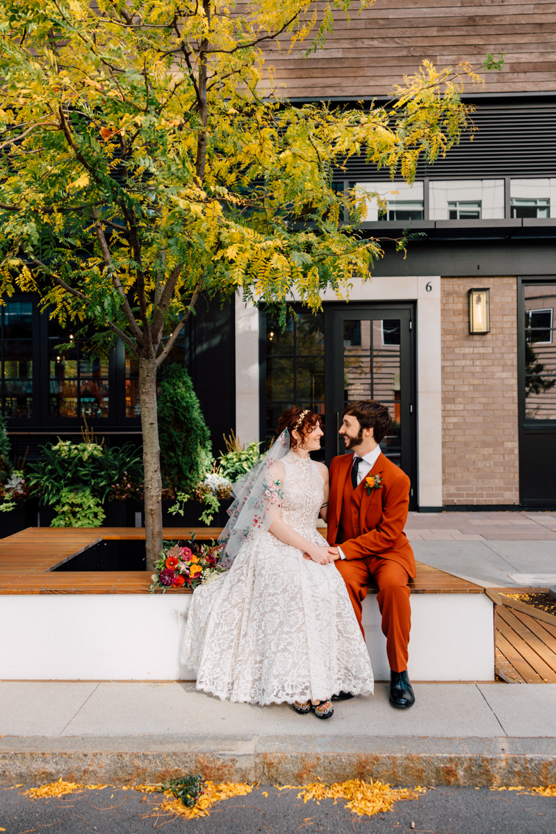 A bride in a lace gown and a groom in a rust-colored suit sit together on a bench surrounded by vibrant fall foliage and greenery.