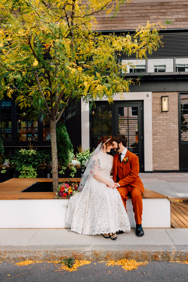 A couple shares a tender moment seated together on an outdoor bench, surrounded by colorful foliage and modern architecture, showcasing their wedding attire and floral bouquet.