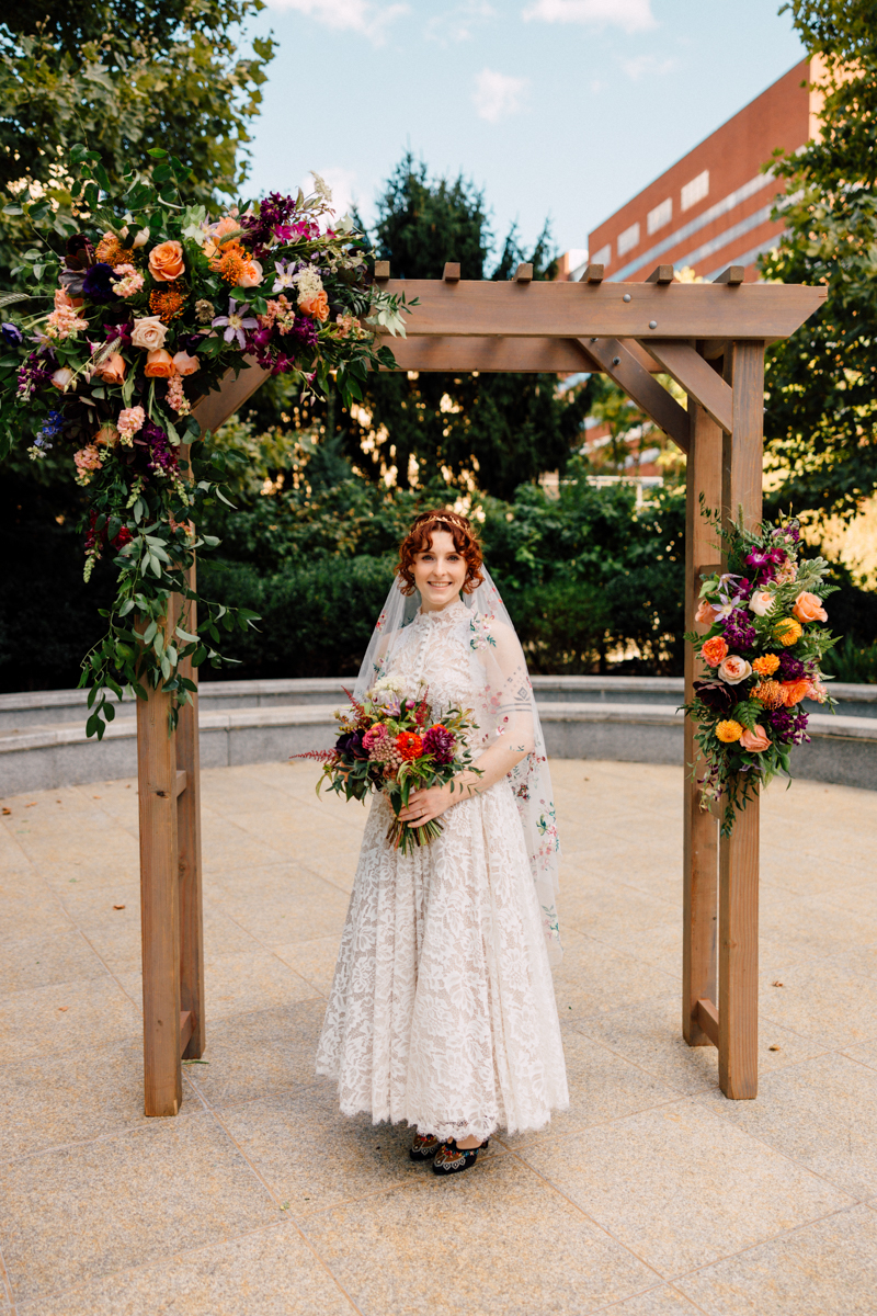 A joyful bride in an intricate lace gown stands under a floral-adorned wooden arch, holding a vibrant bouquet amidst lush greenery.