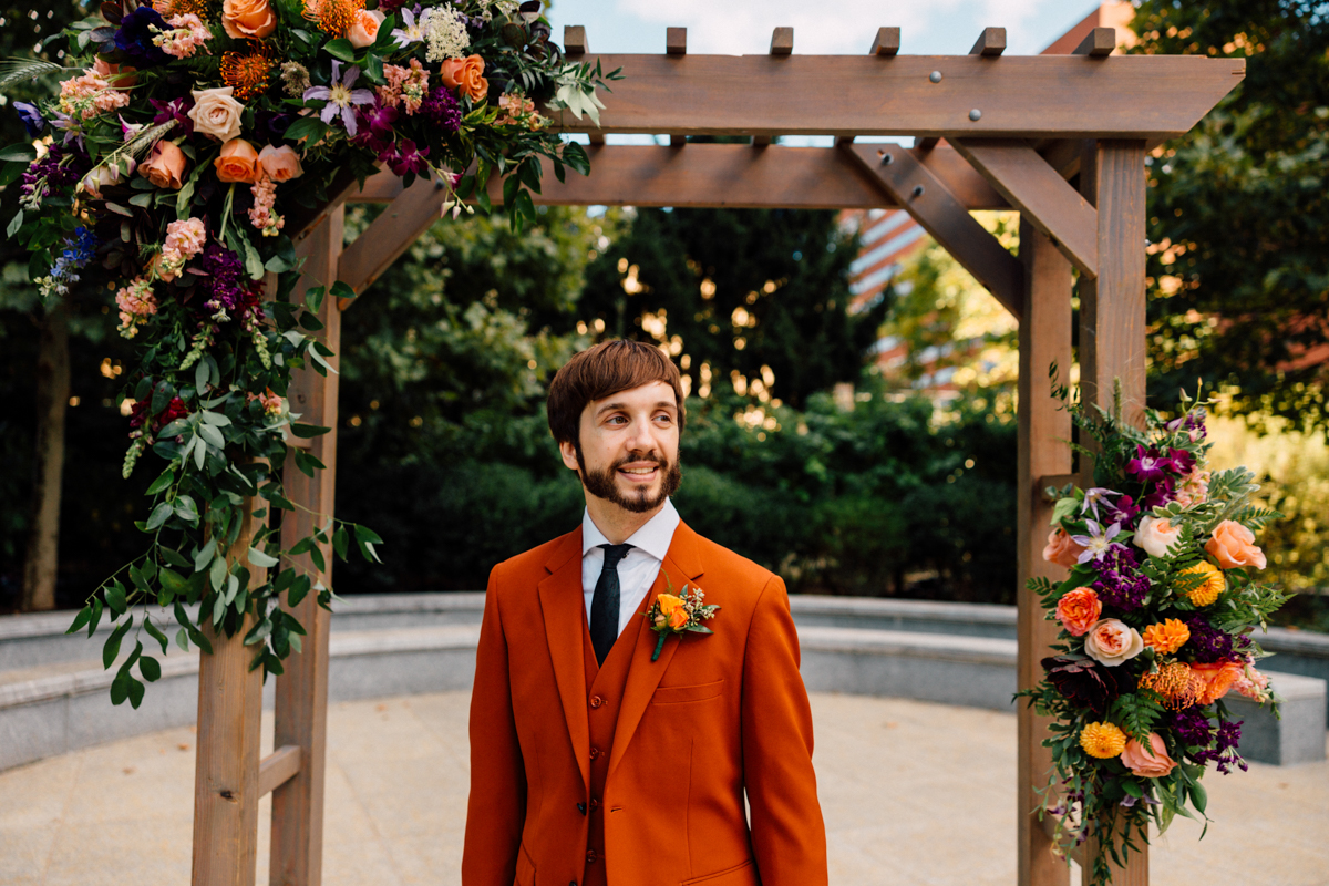 A smiling man in an orange suit stands under a floral archway adorned with vibrant flowers, set in a scenic outdoor location.