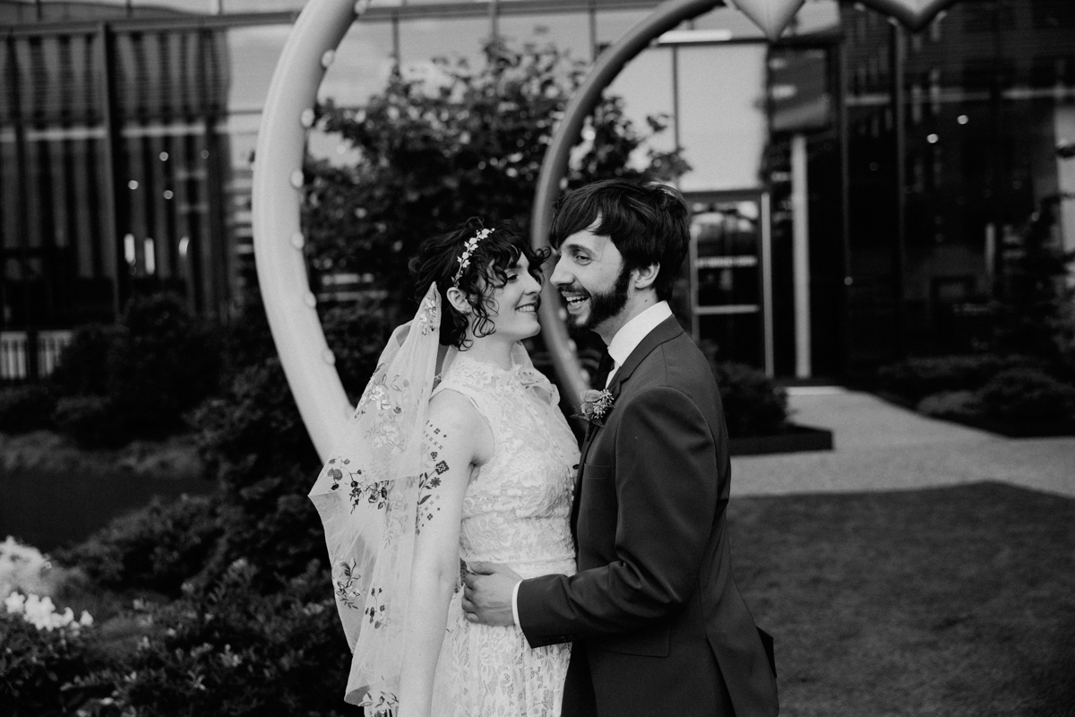 A joyful couple embraces outdoors, sharing smiles in a romantic setting, adorned with greenery and a modern backdrop.