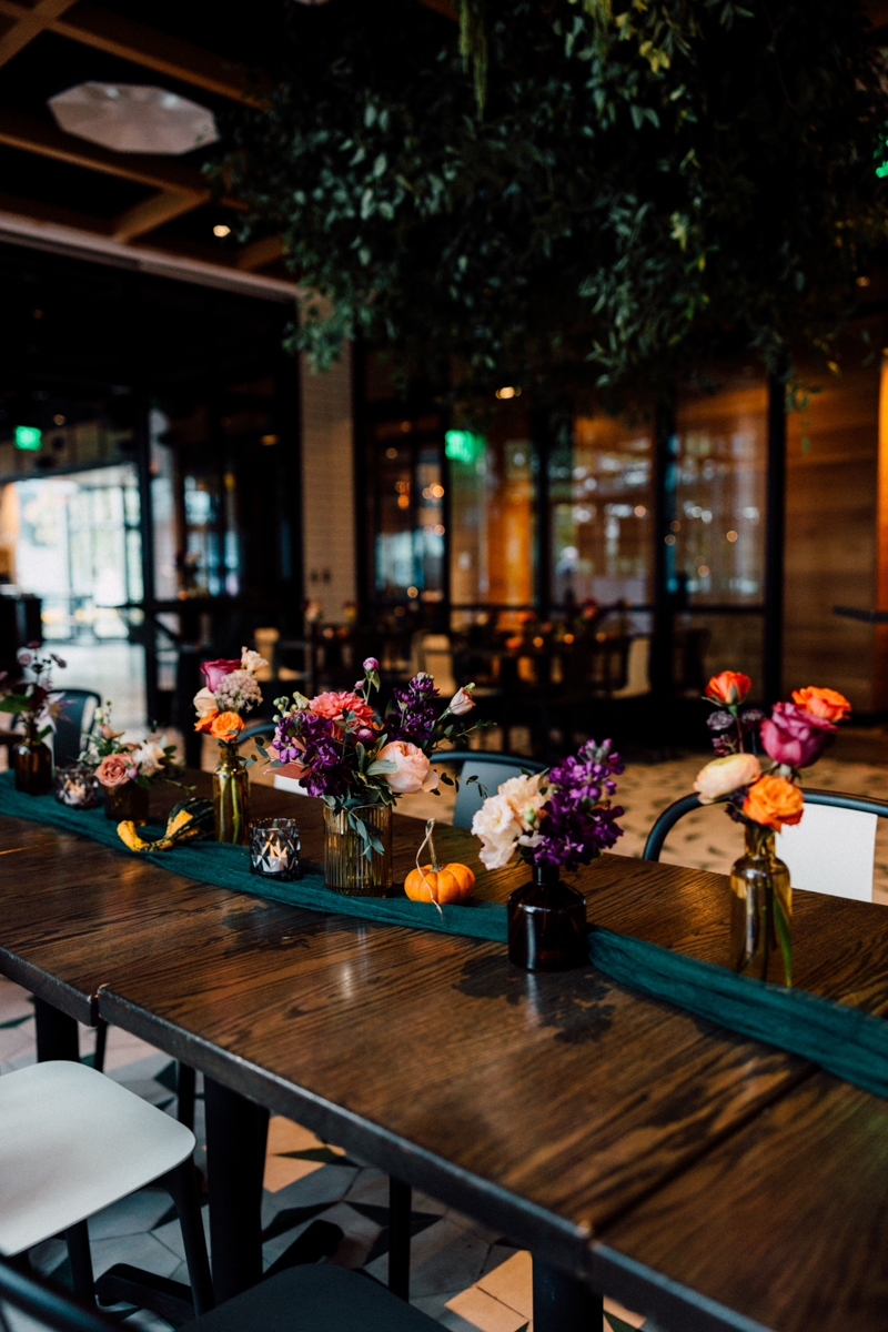 A beautifully arranged table with colorful flowers in glass vases, adorned with a green table runner, candles, and small decorative pumpkins, set in a contemporary restaurant interior.