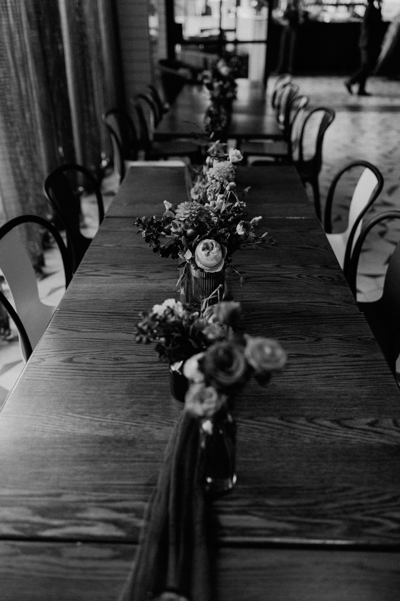 A long wooden table adorned with colorful floral arrangements in glass vases, creating a warm and inviting atmosphere in a stylish interior.
