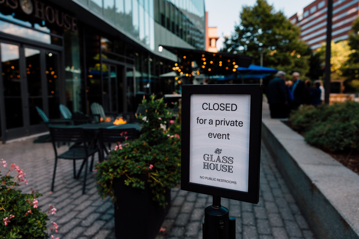 A sign in front of a modern outdoor restaurant area reads, Closed for a private event—Glass House Cambridge Wedding Photography. Patio seating and people are visible in the background under string lights.