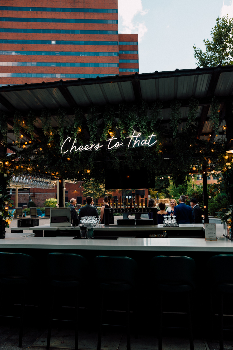 A stylish bar area adorned with a "Cheers to That" neon sign, surrounded by greenery and illuminated by warm lights, with guests mingling in the background.