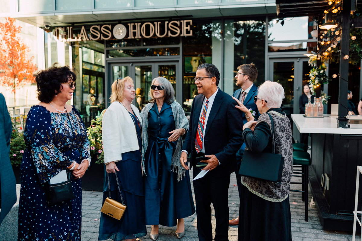 A group of well-dressed adults stand and chat outside a modern building with a sign reading “GLASS HOUSE.” They appear to be attending a social event on a sunny day, perfect for Glass House Cambridge wedding photography.