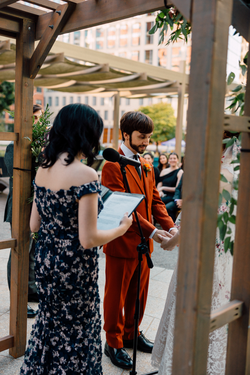 A groom in an orange suit exchanges vows with the bride during an outdoor wedding ceremony, while a woman officiates and guests look on in the background.