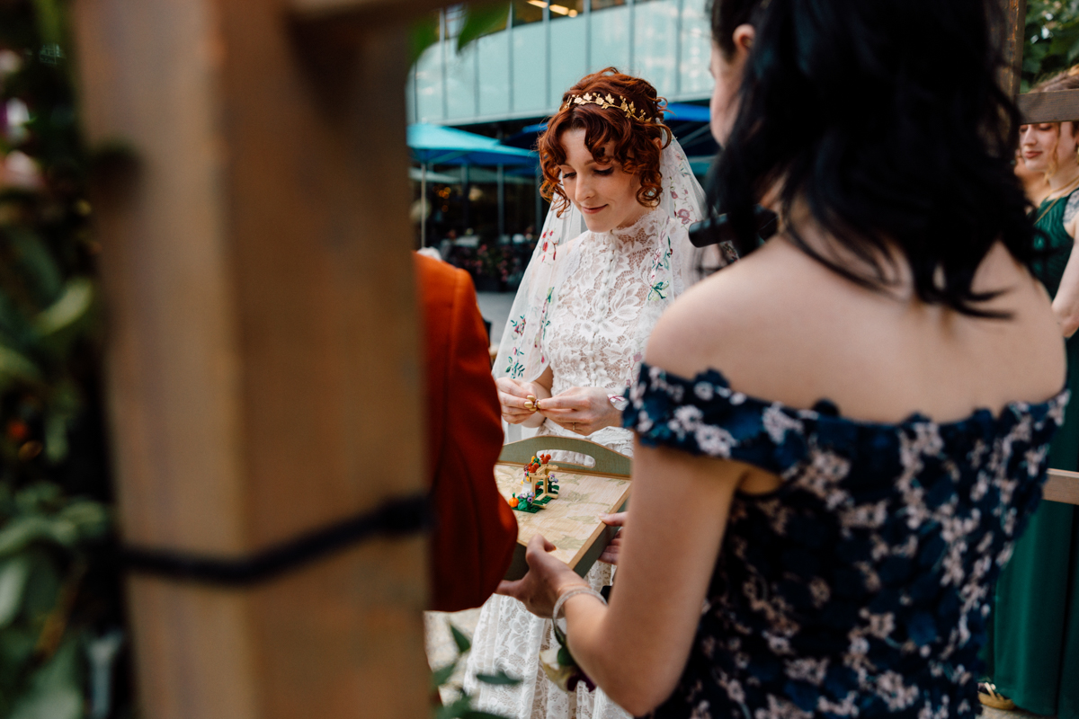 A bride in a floral lace dress and veil prepares for a ceremonial exchange, holding a ring while surrounded by friends and colorful decorations.