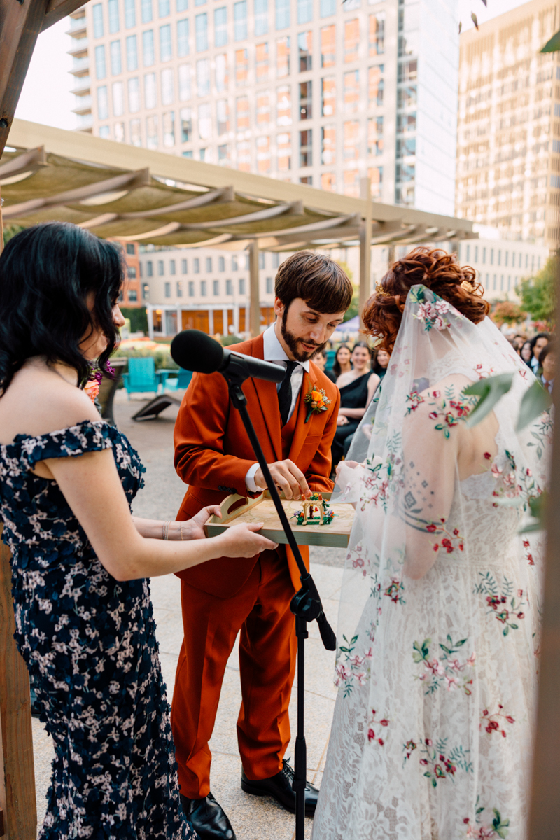 A bride and groom participate in a wedding ceremony, with the groom in an orange suit and the bride in a floral lace gown, exchanging a small cake while a guest officiates.