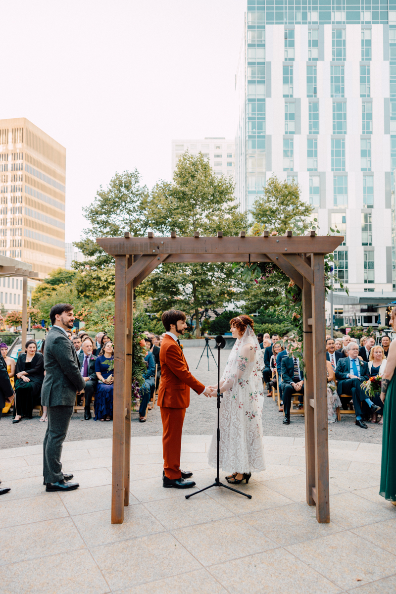 A couple exchanging vows under a wooden arbor, surrounded by guests in an outdoor setting with modern buildings in the background.