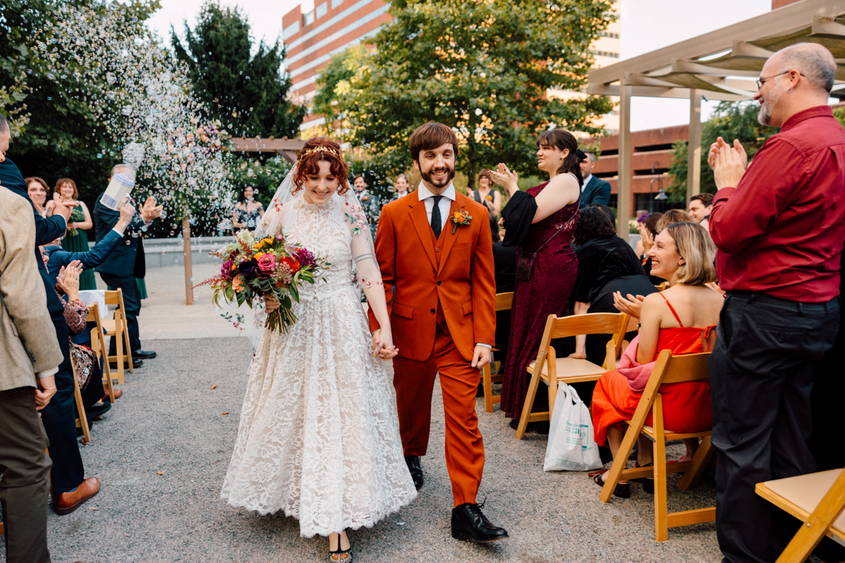 A happy couple walks hand in hand, surrounded by guests celebrating their marriage with confetti, in a beautifully decorated outdoor setting.