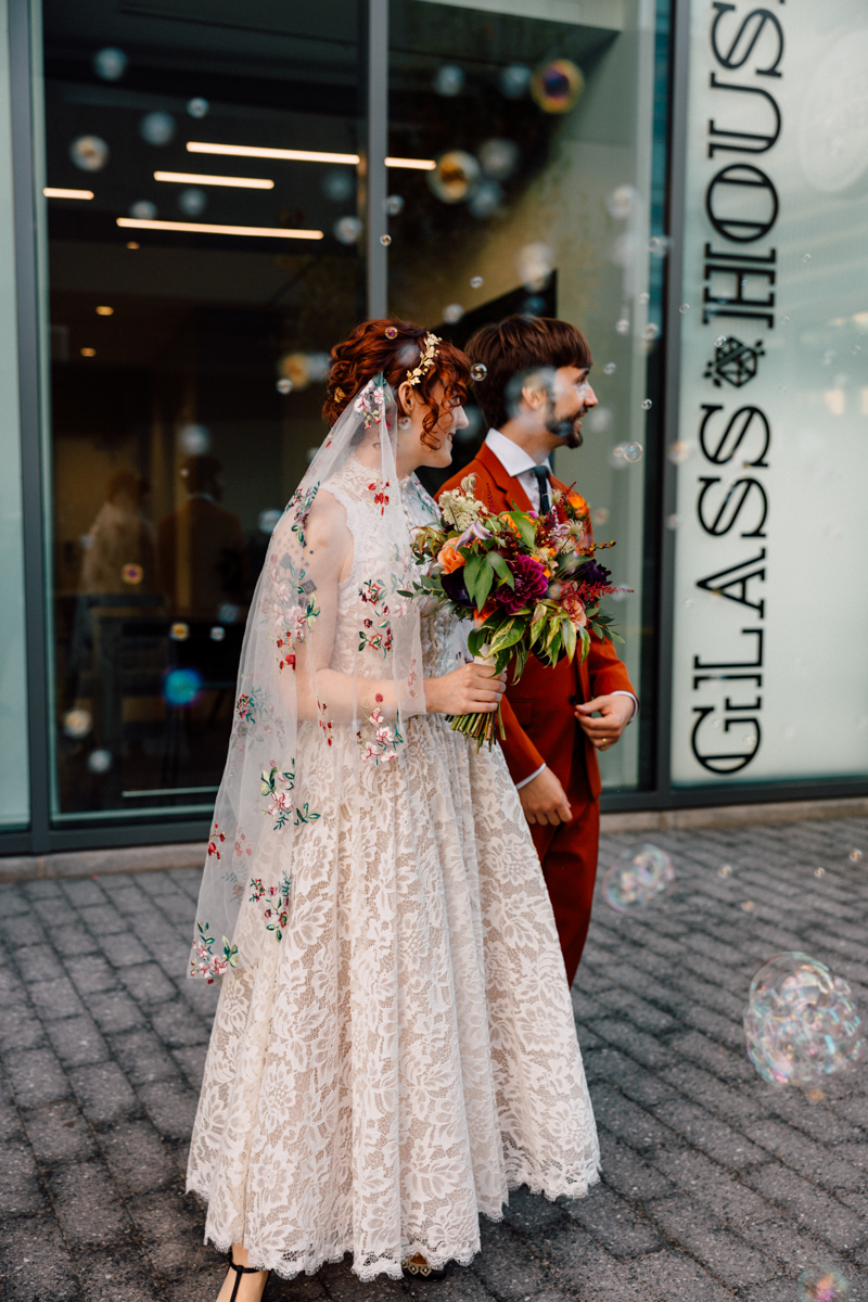 A couple joyfully walks outside in wedding attire, surrounded by floating bubbles, with the bride in a floral lace dress and veil, and the groom in a striking red suit.