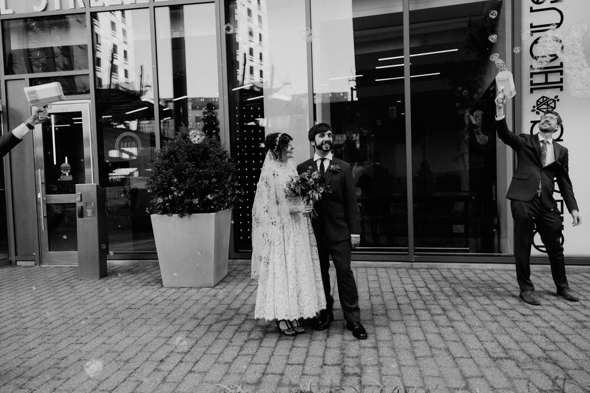 A joyful couple poses for a photo outside a venue while a guest sprays bubbles in the background.