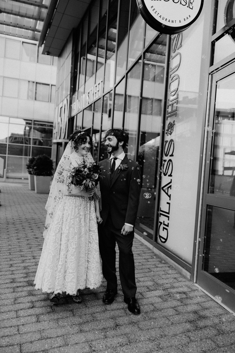A joyful couple in wedding attire stands outside a modern venue, surrounded by bubbles, exuding love and happiness.