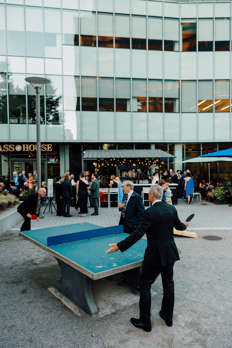 Two men in formal attire engage in a lively game of ping pong outside, surrounded by a festive gathering and modern architecture.