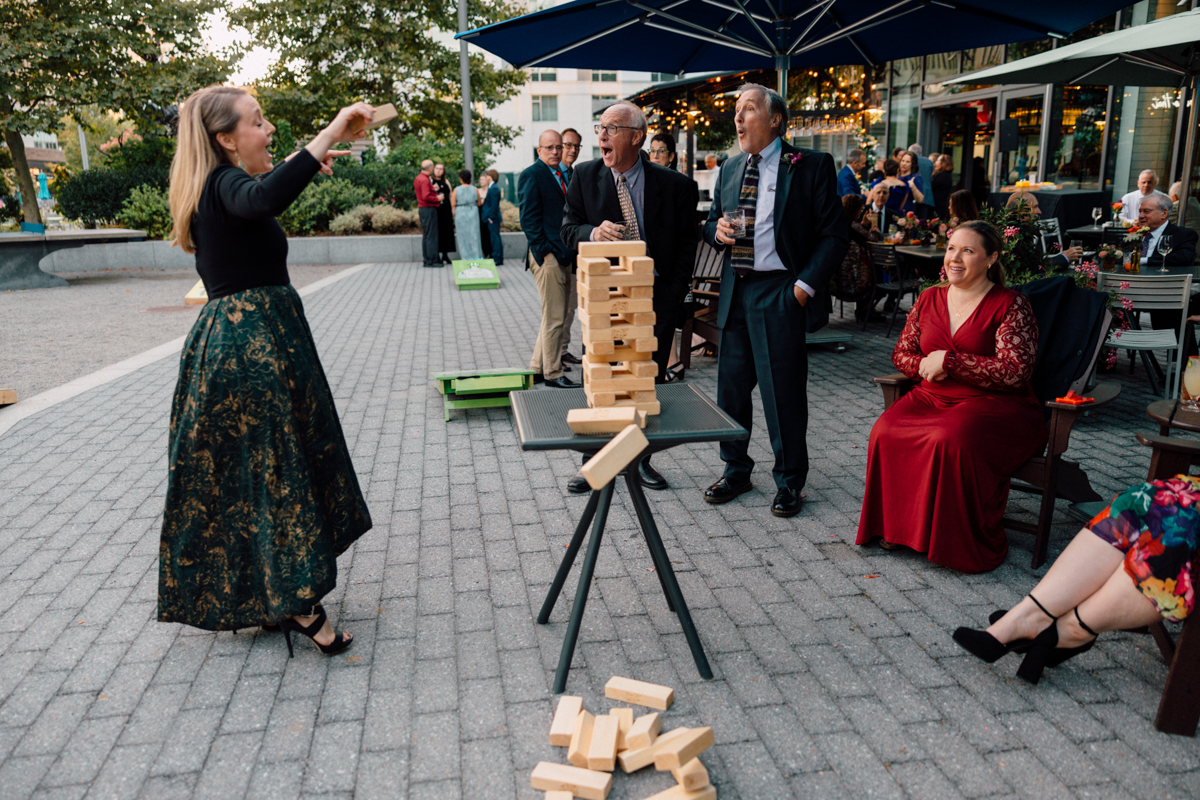 A woman playfully removes a block from a Jenga tower while onlookers watch in amusement at an outdoor event.