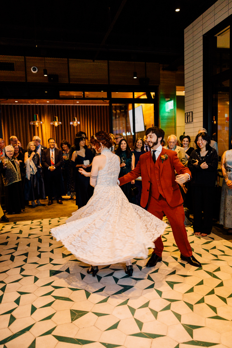 A couple dances joyfully at their wedding reception, surrounded by smiling guests in an elegant venue with patterned flooring.