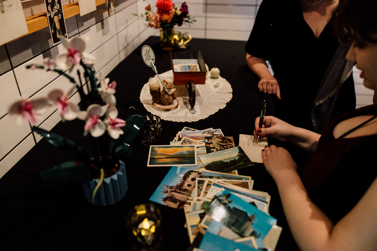 A woman writes on a postcard at a black table adorned with vintage postcards, decorative flowers, and a small figurine, capturing a nostalgic atmosphere.