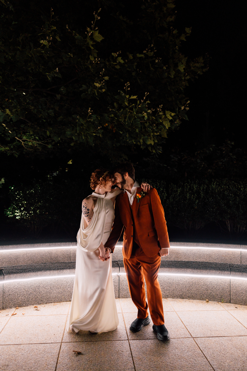A couple in an intimate moment, dressed in wedding attire, standing close together under nighttime lights, with a backdrop of greenery.
