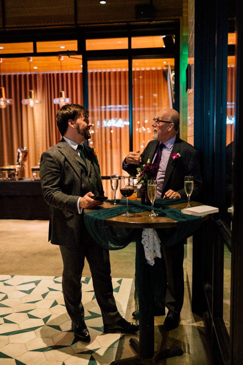 Two men engage in a lively conversation at a stylish event, surrounded by drinks and elegant decor.