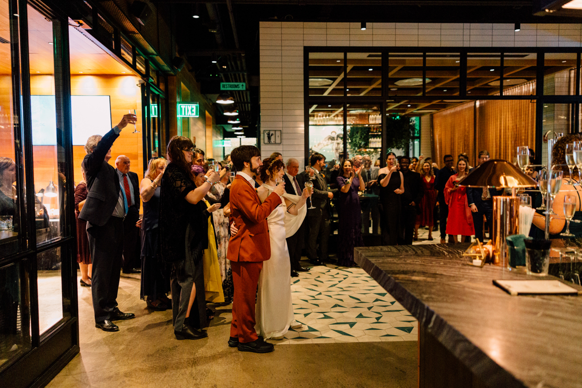 A joyful crowd raising glasses in a toast during a celebration, featuring a couple in formal attire at the center, surrounded by friends and family in an elegant venue.