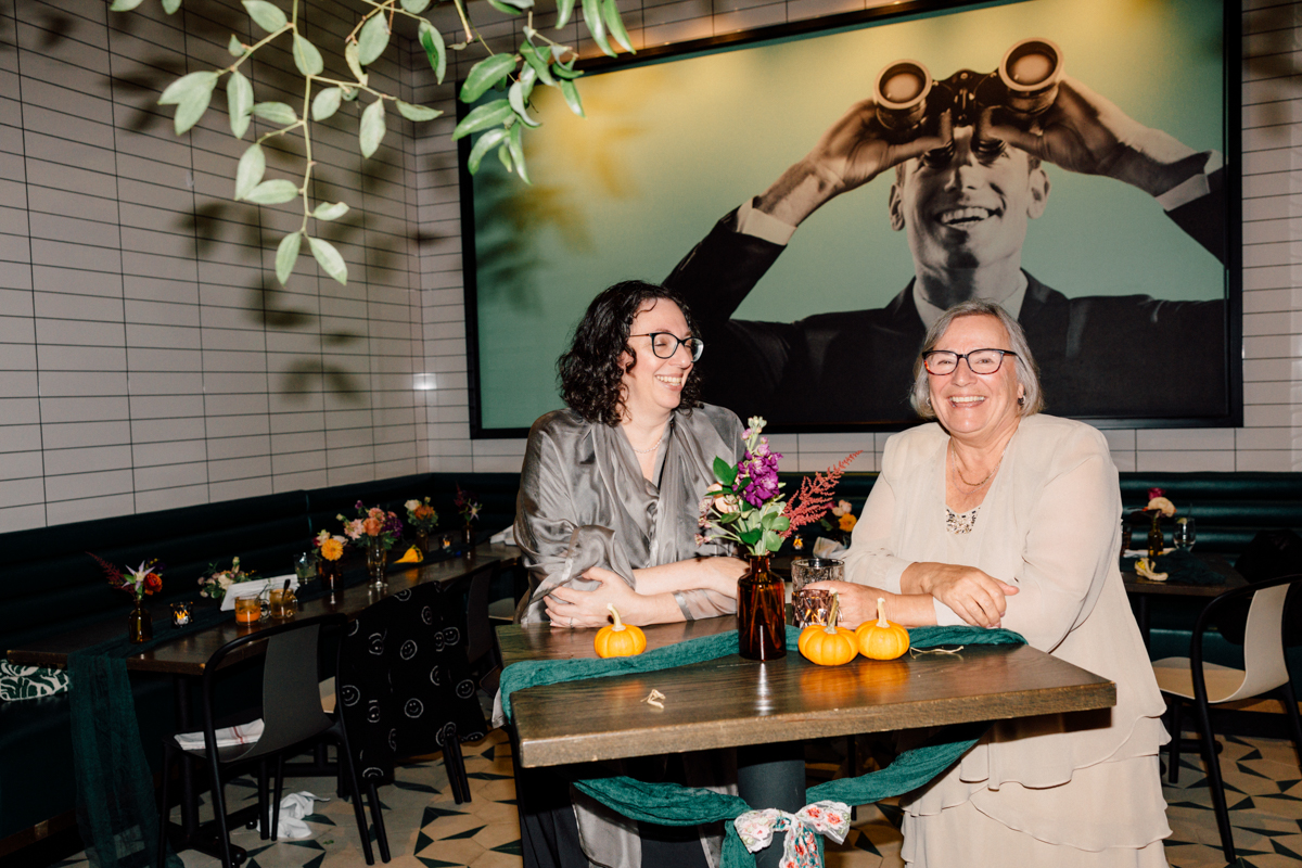 Two women laughing and engaging at a table with flowers and pumpkins in a chic café setting, featuring a large decorative mural in the background.