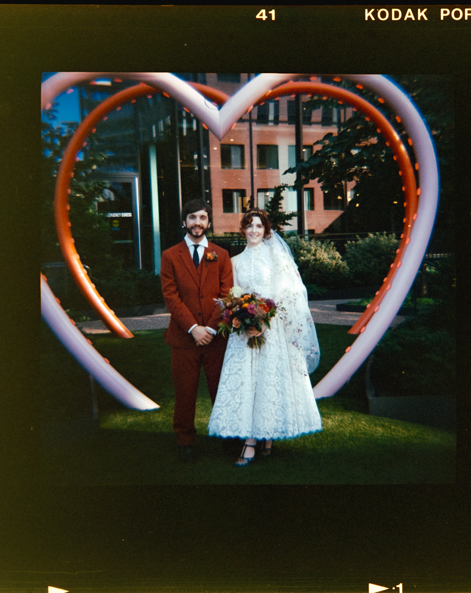 A couple stands together in front of a heart-shaped light installation, showcasing the groom in a red suit and the bride in a white lace dress, holding a colorful bouquet.
