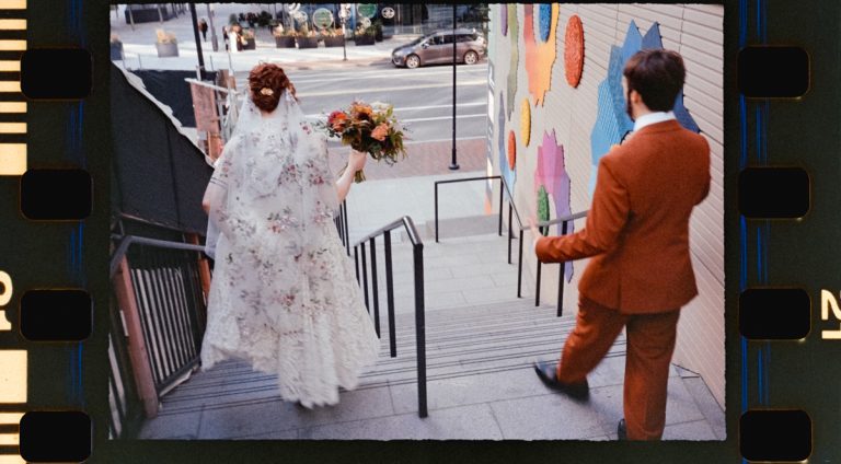 A bride in a floral lace dress and veil holds a bouquet while walking down the stairs, accompanied by a man in a rust-colored suit. The backdrop features colorful wall art. Photo taken on 35mm film