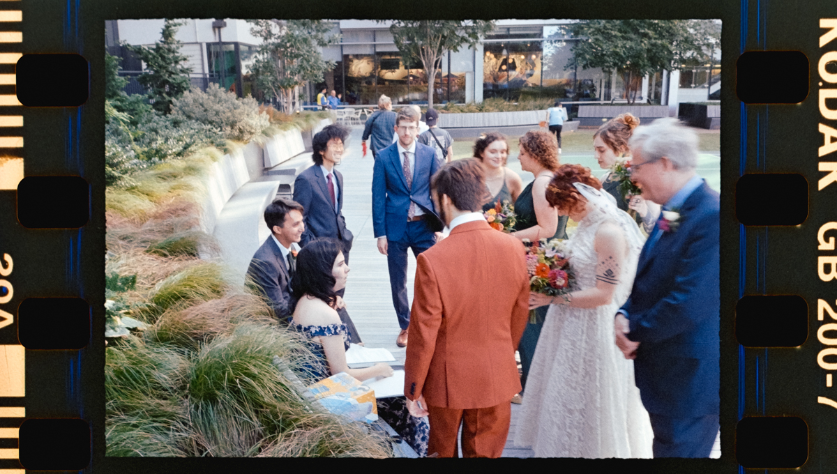 A group of elegantly dressed individuals gather in a lush garden, with bride and groom at the center, surrounded by friends and family, amidst greenery and stylish architecture.