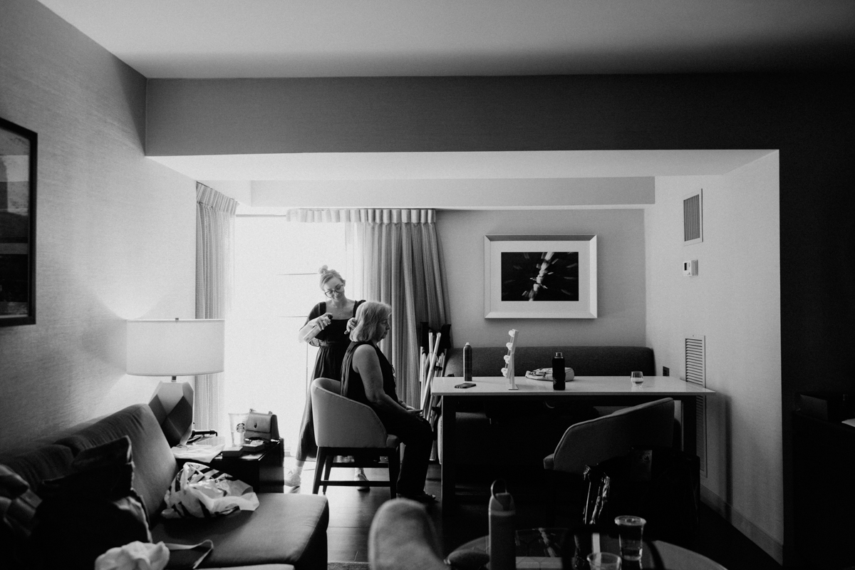 A hairstylist does hair in a hotel room while a client sits patiently in a chair, surrounded by personal items and a bright window.