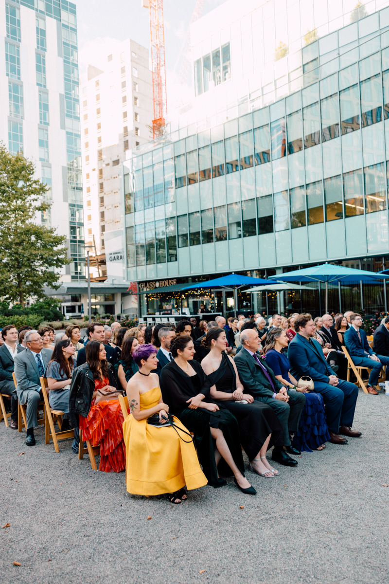 A diverse audience seated outdoors, dressed in formal attire for a wedding ceremony surrounded by modern buildings and greenery.