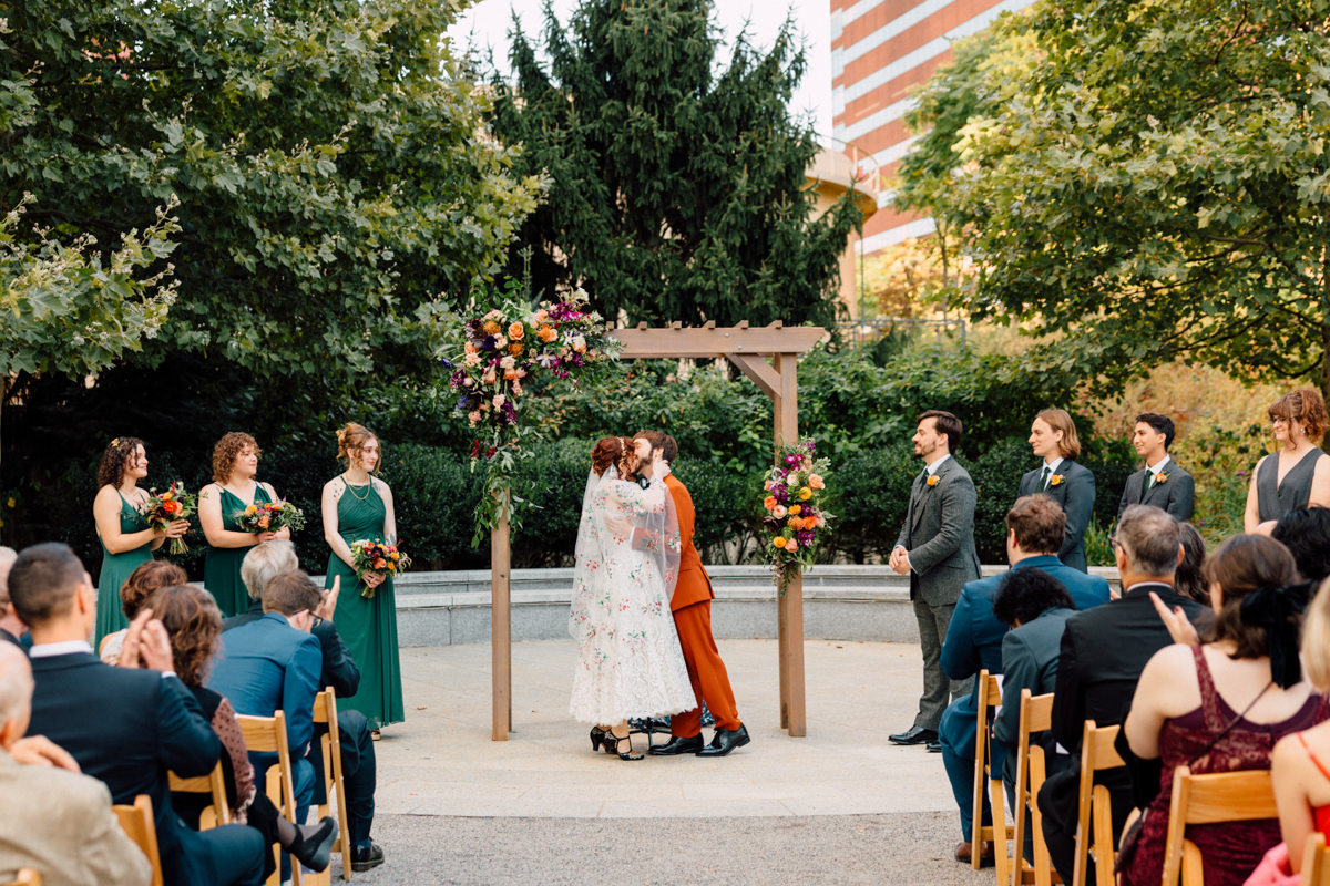 A couple embraces during their wedding ceremony under a floral arch, surrounded by bridal party members and guests.