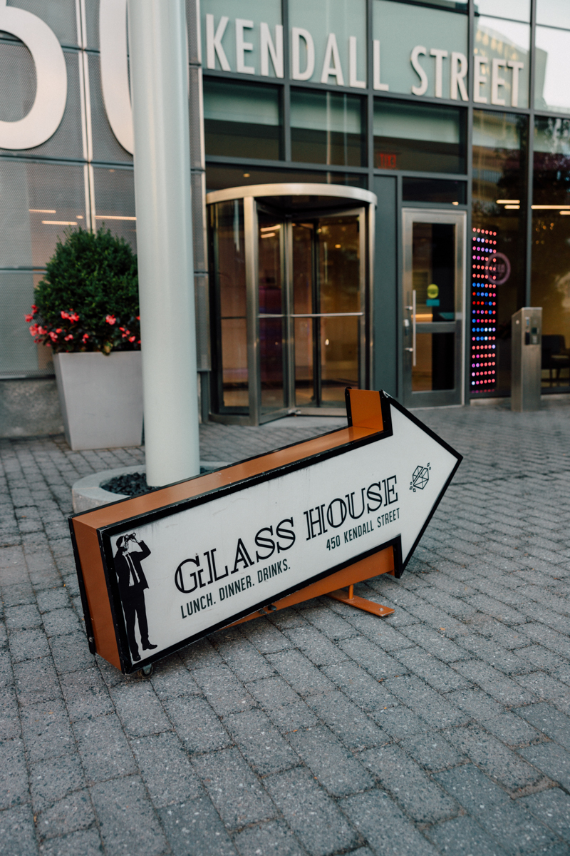 A large arrow-shaped sign for Glass House Cambridge sits on a sidewalk, pointing right. The restaurant offers lunch, dinner, and drinks at 450 Kendall Street. Perfect spot for Glass House Cambridge wedding photography, with a glass building entrance behind.