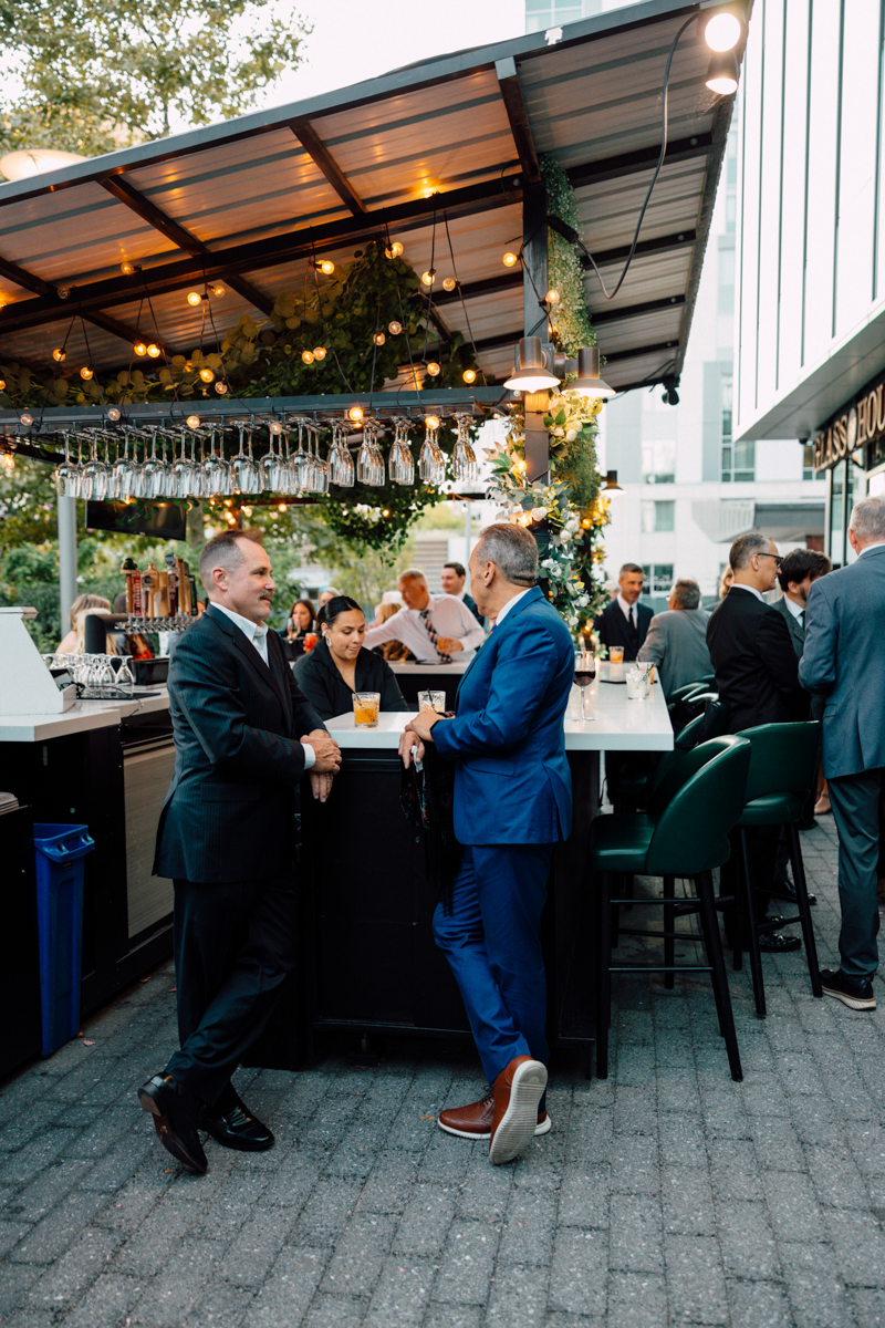 Two men in suits conversing at an outdoor bar with others enjoying drinks in the background.