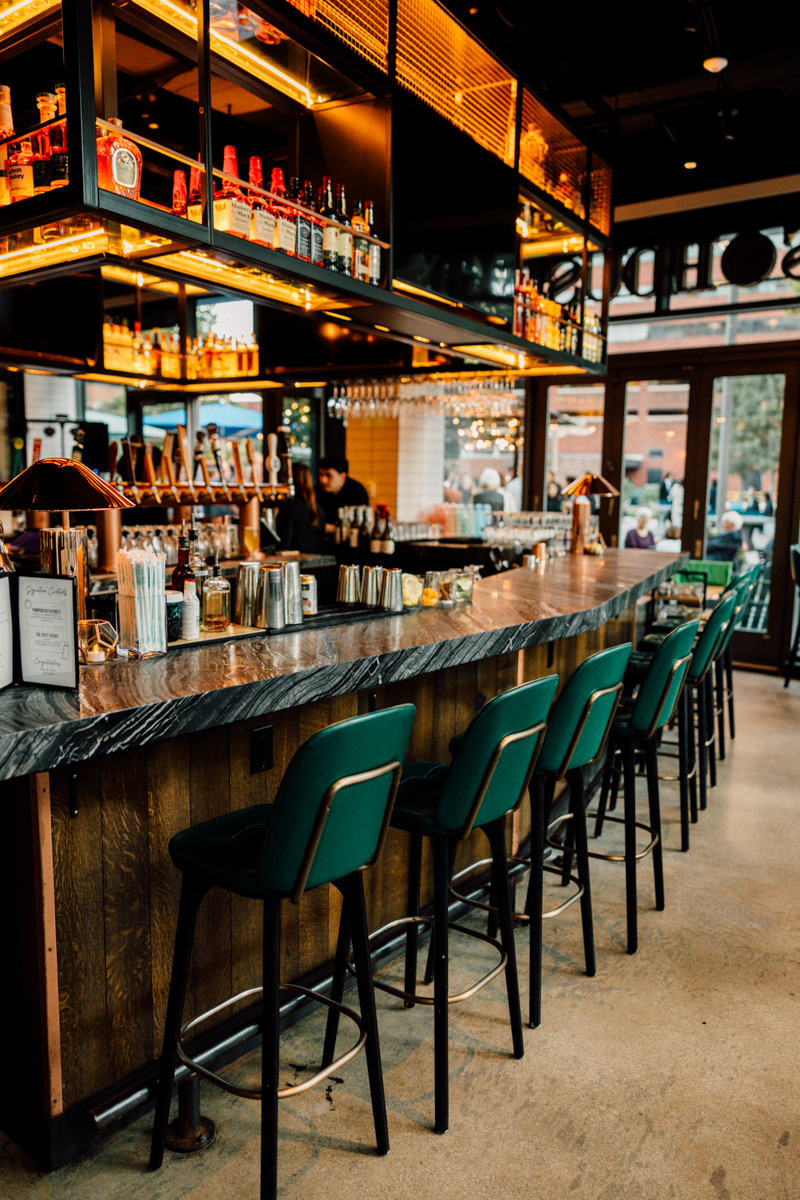 A modern bar featuring a sleek marble countertop, illuminated shelves of liquor bottles, green bar stools, and a warm, inviting atmosphere.