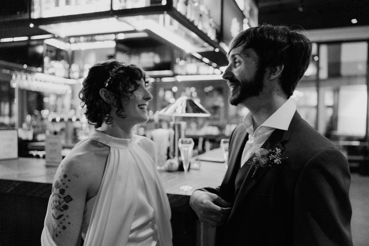 A joyful couple dressed in formal attire stand facing each other and smiling in a softly lit bar, shelves of glasses and bottles behind them. Captured in black and white, this moment embodies the elegance of Glass House Cambridge Wedding Photography.