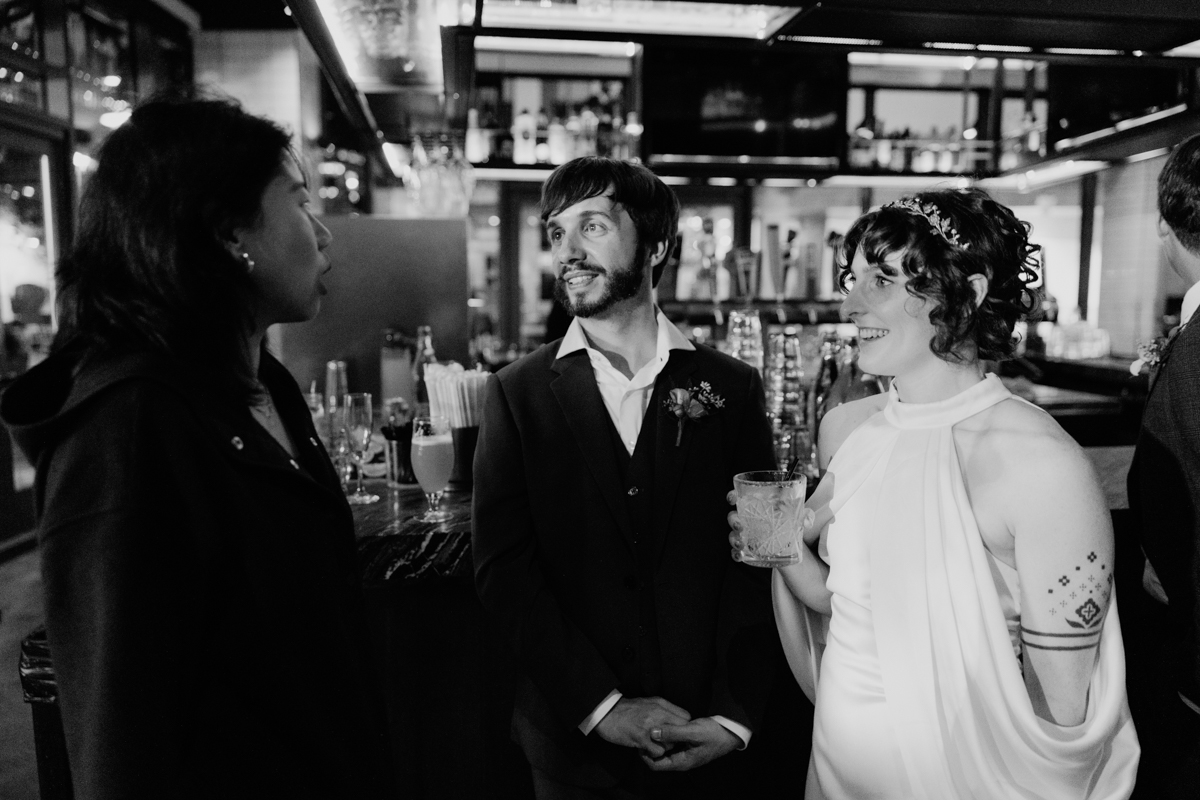 Three friends chatting at a bar, with one holding a cocktail, while the others listen attentively in a lively atmosphere.