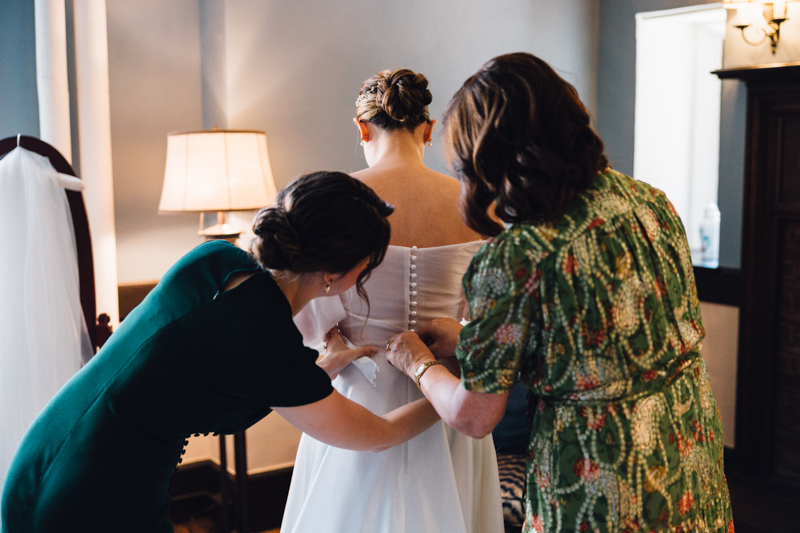 Two women help a bride button and tie the back of her dress at the Pavilion at Grace Wedding. The bride stands with her back to the camera in a softly lit room, with a lamp and some furniture visible in the background.
