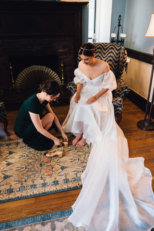 A bride in a white dress sits on a chair at the Pavilion at Grace Wedding, while another woman kneels to help her put on gold shoes in a warmly lit room with wooden floors and a patterned rug.