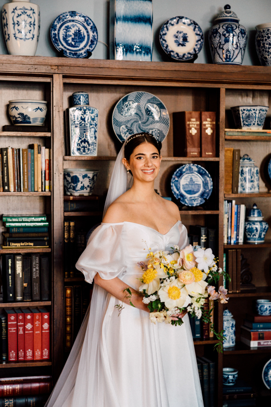 A bride in an off-the-shoulder white gown stands before bookshelves lined with blue-and-white porcelain at her Pavilion at Grace Wedding, holding a bouquet of yellow, white, and pink flowers and smiling at the camera.
