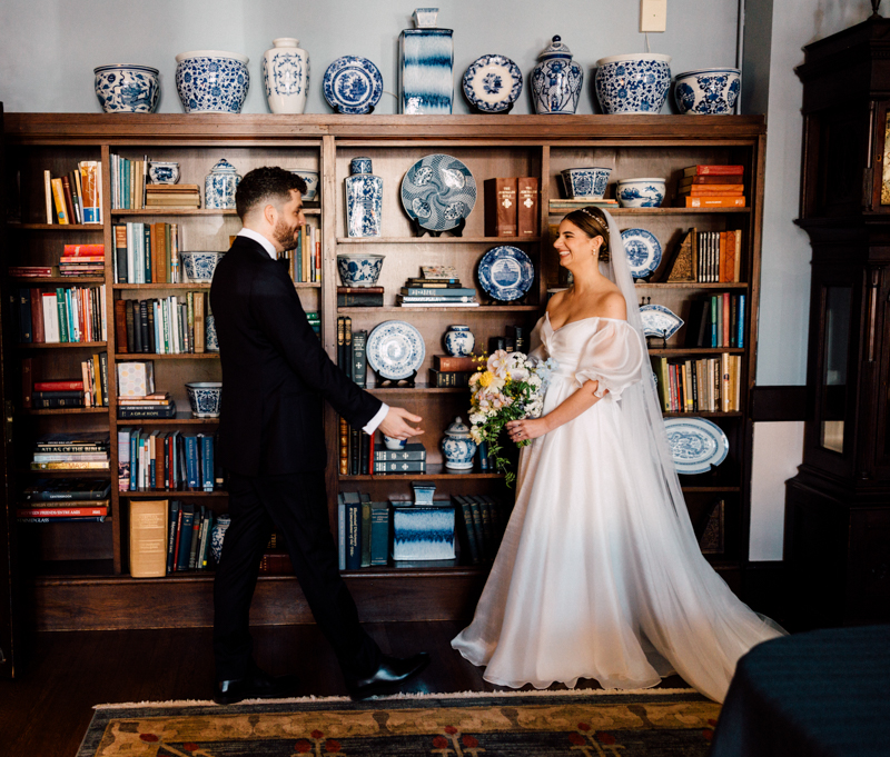 A groom in a dark suit smiles and extends his arms toward a bride in a white gown holding a bouquet. They stand in front of a bookshelf decorated with blue and white porcelain, capturing their Pavilion at Grace Wedding moment.