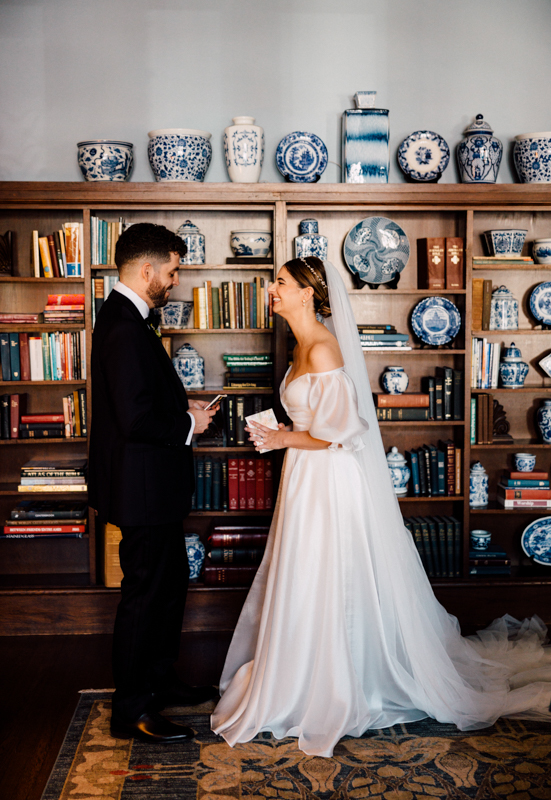 A bride in a white dress and veil smiles at her groom in a black suit at the Pavilion at Grace Wedding. They stand facing each other in front of bookshelves adorned with blue and white porcelain vases and plates.