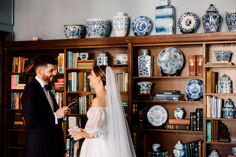 A bride and groom stand smiling in front of bookshelves adorned with blue and white porcelain vases at their Pavilion at Grace Wedding, as the groom holds a phone and the bride raises her glass, both dressed in elegant wedding attire.
